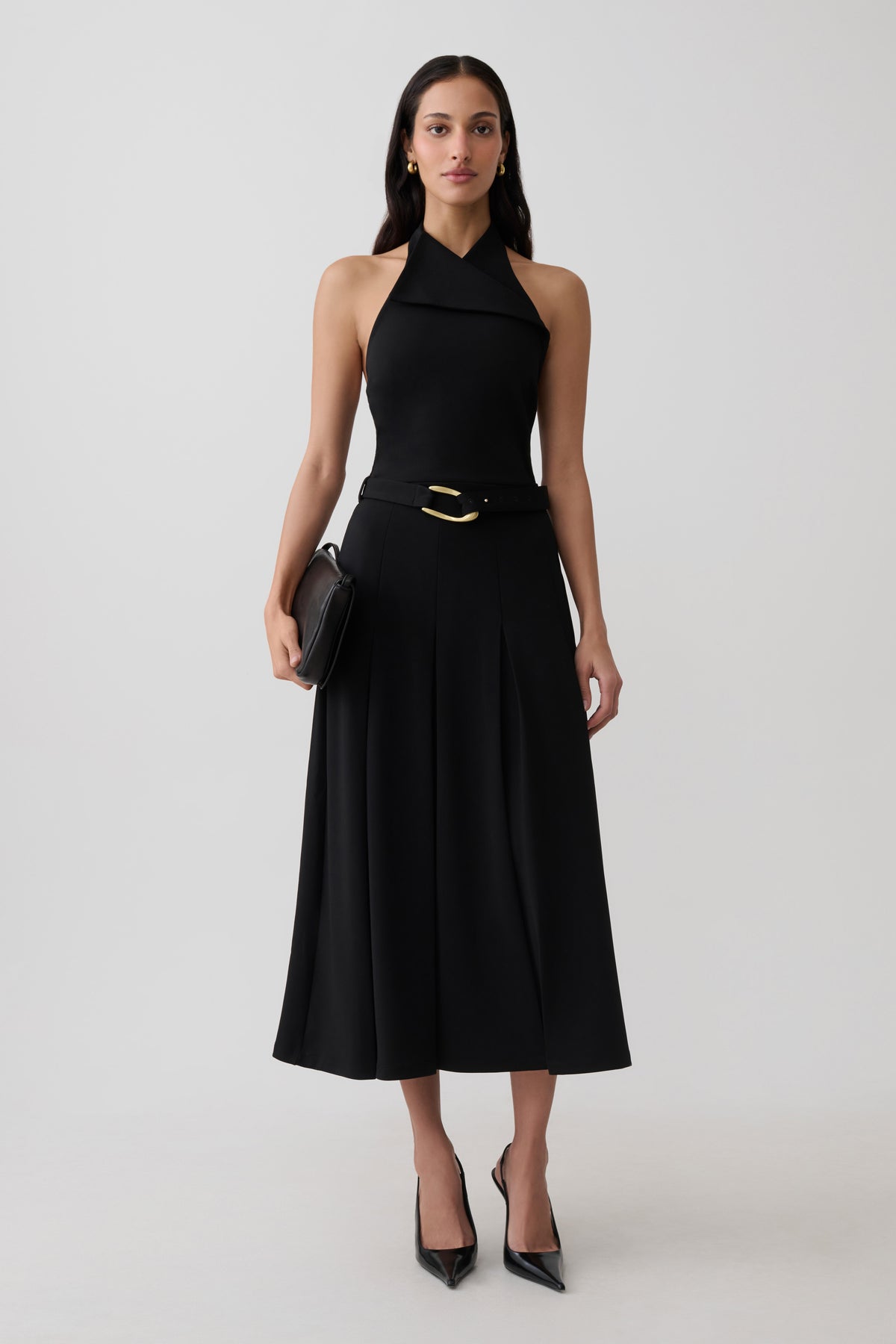 A woman modeling a sleeveless black midi dress with a halter-style neckline, a matching black belt with a gold buckle, black pointed-toe heels, and a small black clutch, photographed against a light gray studio background.