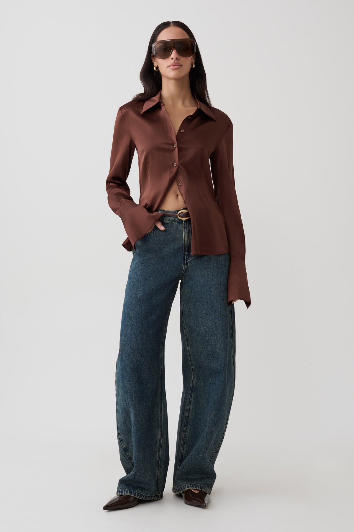A model stands against a light gray studio background wearing a brown long-sleeve button-up shirt, blue wide-leg jeans, dark sunglasses, and dark pointed shoes.