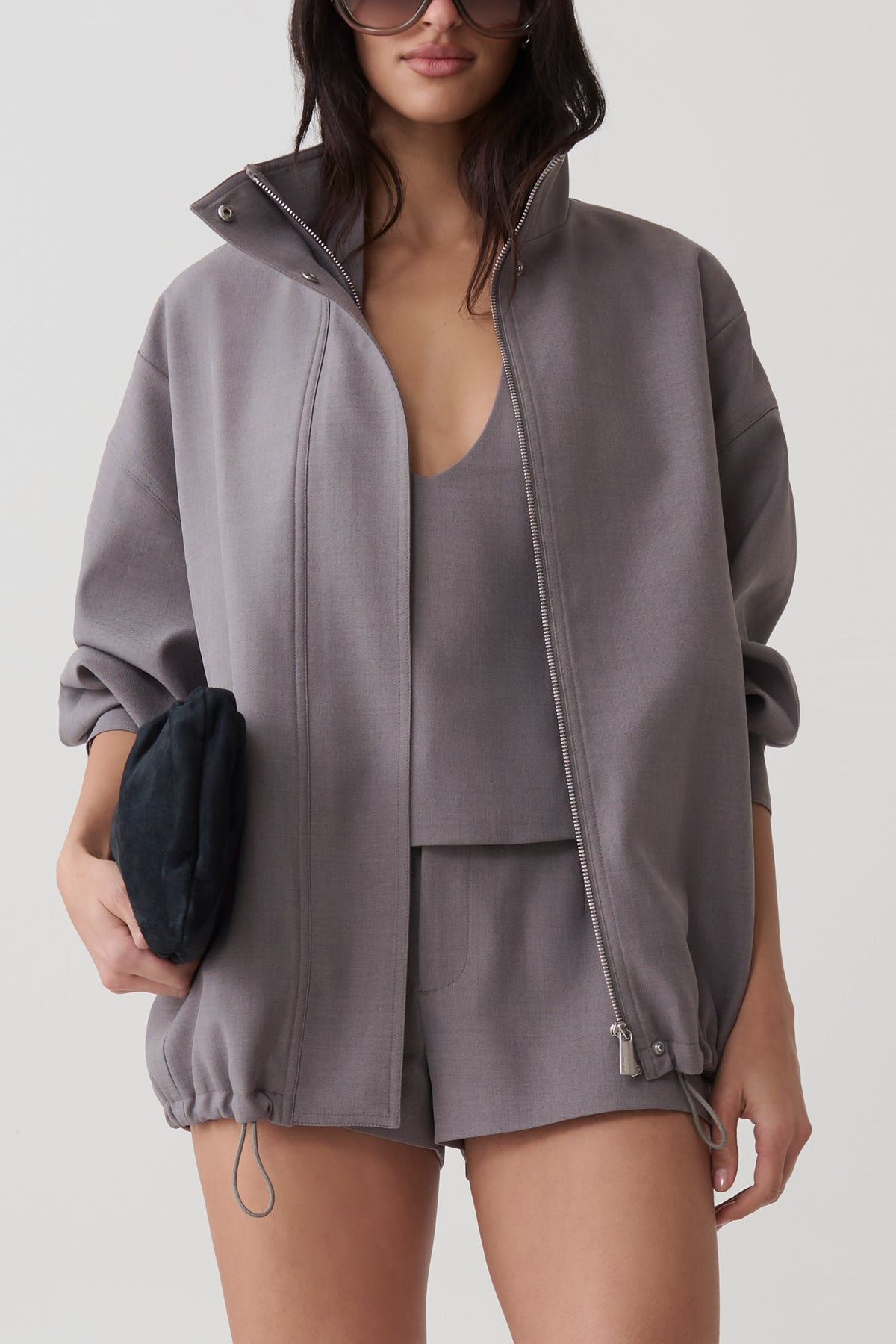 A woman models an oversized gray zip-up jacket with a stand collar, paired with matching shorts and a matching camisole. She holds a small black clutch against a clean, light gray studio background.