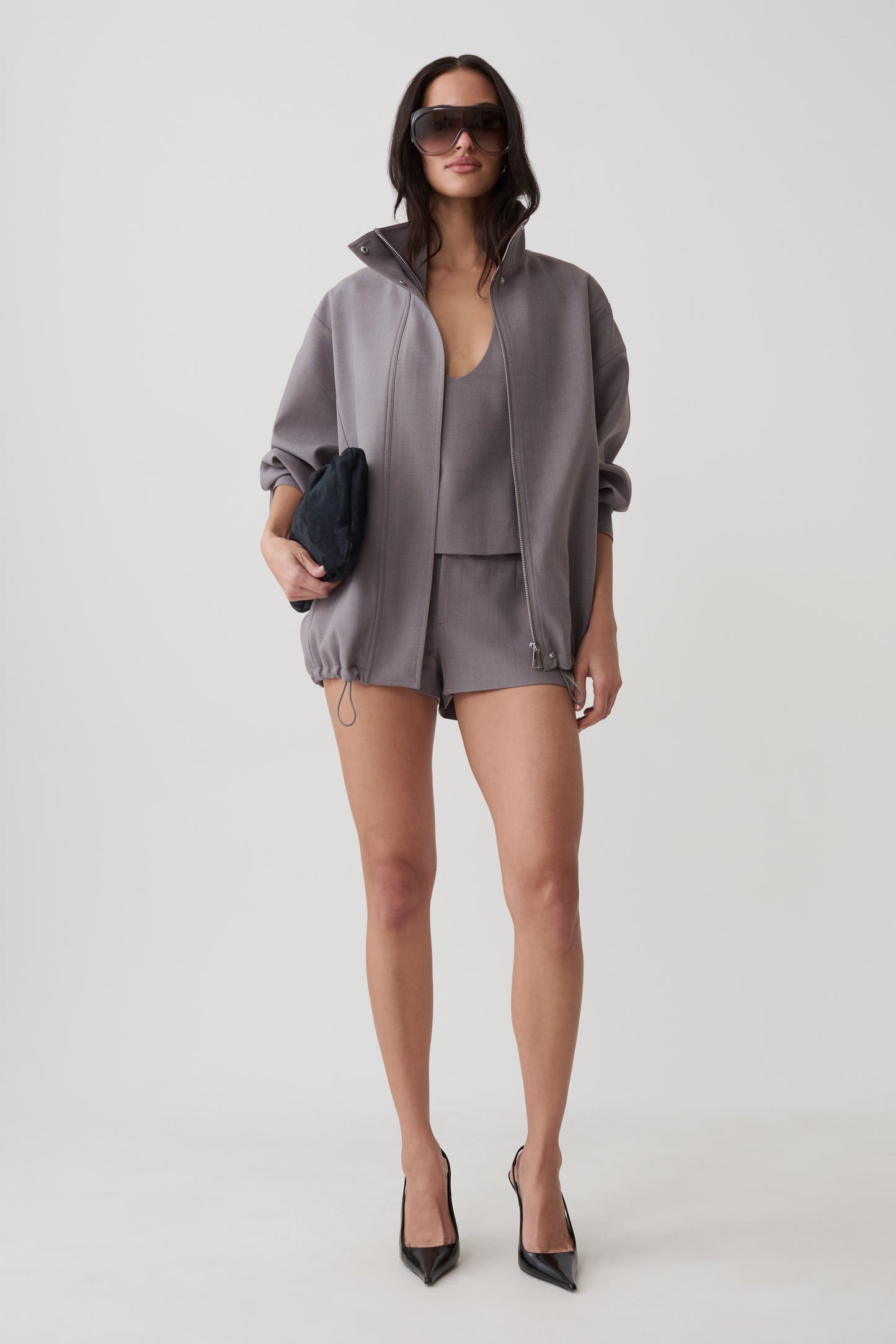 A model stands against a light gray studio background wearing a coordinated gray outfit consisting of an oversized zip-front jacket, matching top, and shorts. She is also wearing black pointed heels and holding a small dark clutch.