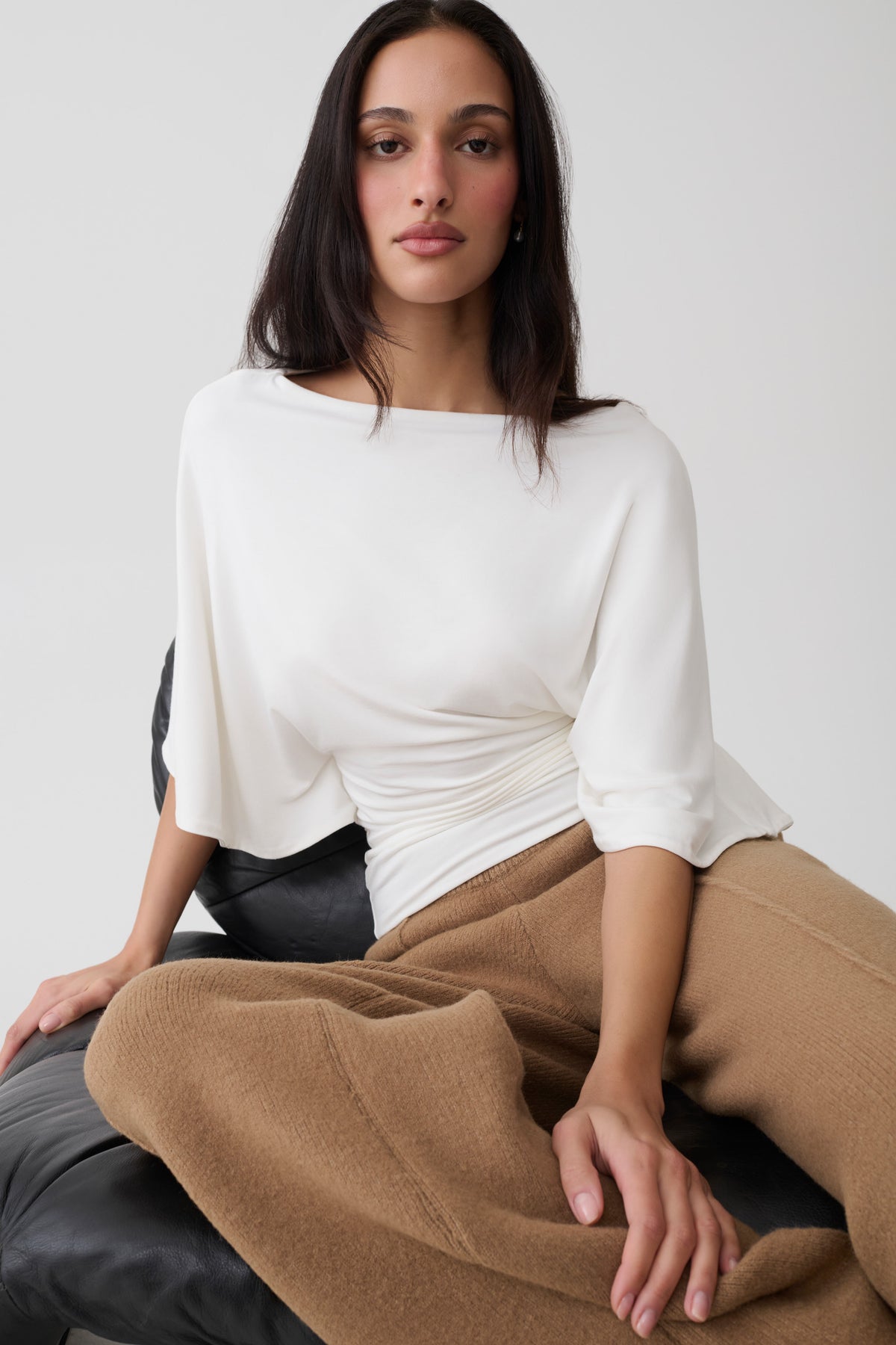 A woman wearing a white draped three-quarter sleeve top and tan wide-leg trousers, seated against a light gray background.