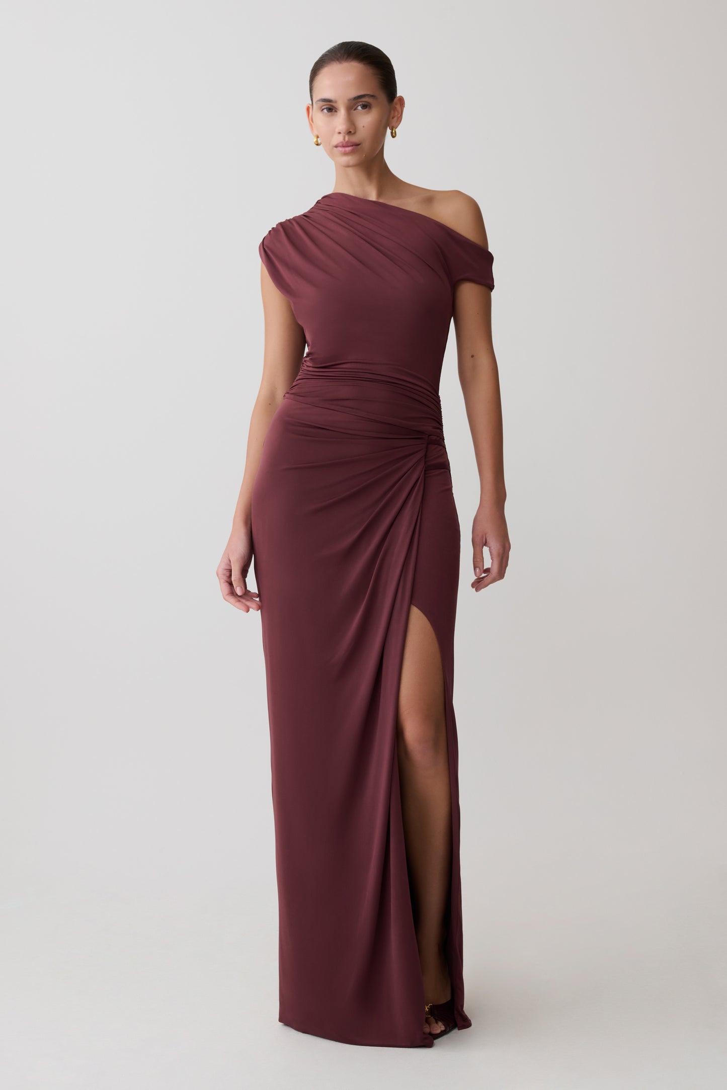 Woman wearing a burgundy off-shoulder gown with a slit on a light gray background
