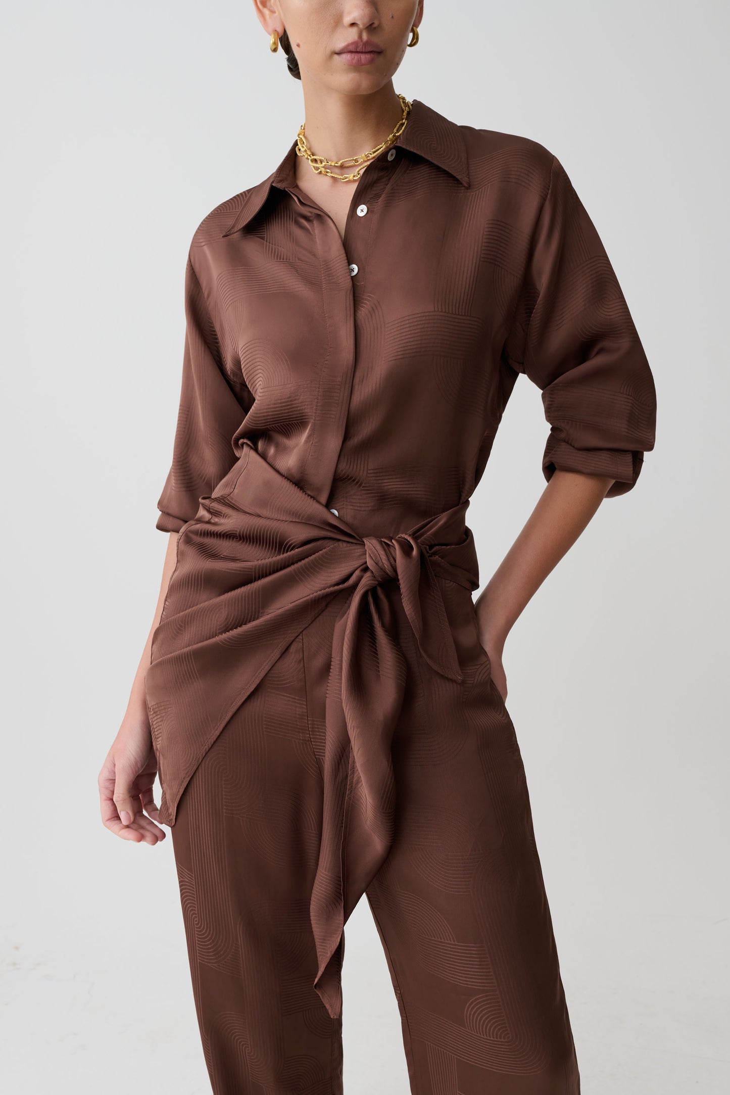 Person wearing a brown shirt and pants with a tied waist on a plain background