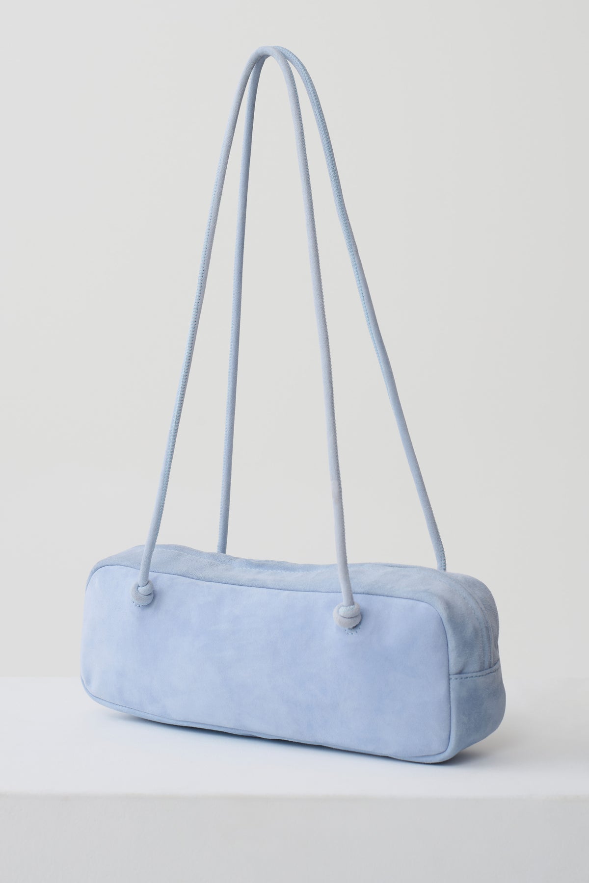 A light blue suede shoulder bag with a long, slender cylindrical body and matching shoulder straps, shown on a clean white studio background.