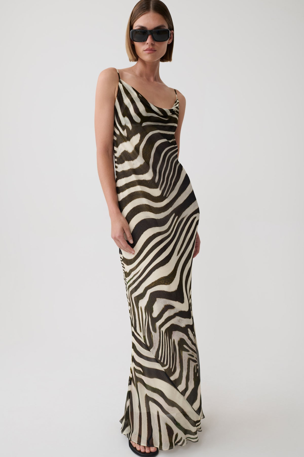 A woman modeling a sleeveless, floor-length zebra-print dress with a draped neckline and thin straps, photographed against a plain light gray studio background.