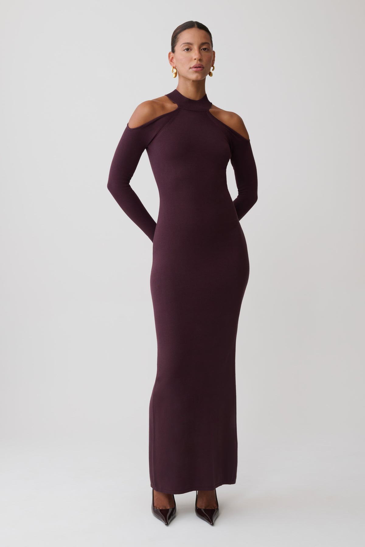 A woman modeling a long-sleeve, high-neck, bodycon maxi dress in a deep plum purple color, styled with pointed-toe heels against a light gray studio background.