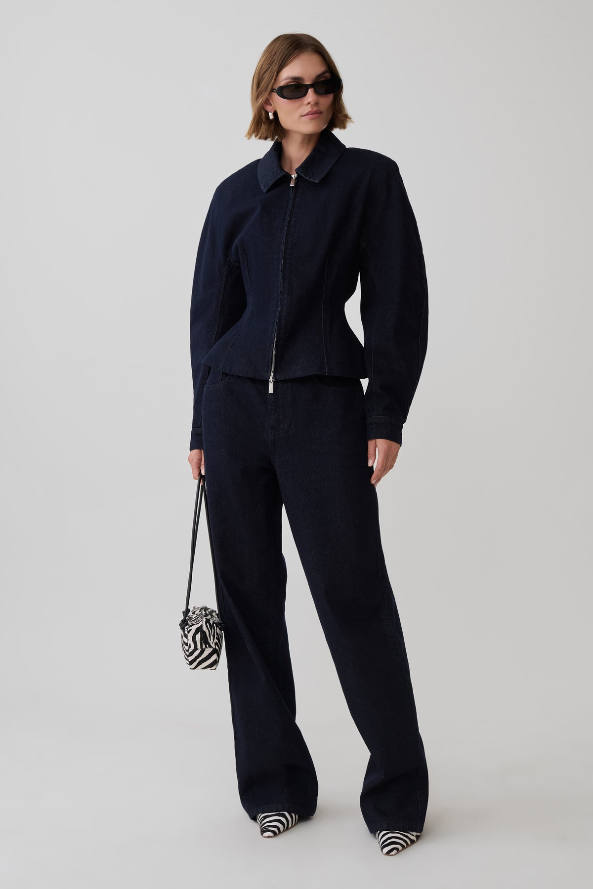 A model stands against a light gray studio background wearing a matching dark navy blue zip-up jacket and wide-leg trousers set, styled with black sunglasses, a small black-and-white zebra-patterned handbag, and black-and-white striped shoes.