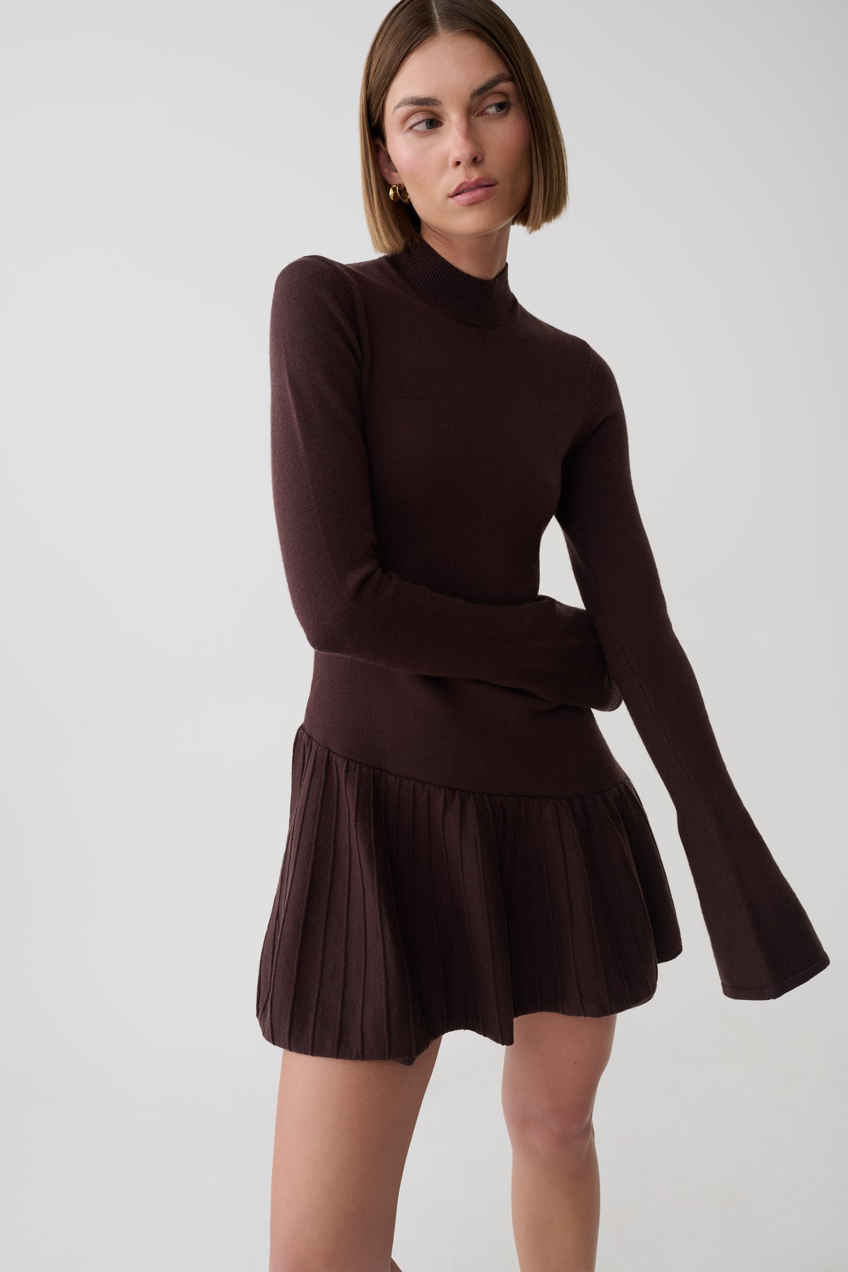 A woman modeling a dark brown ribbed knit mock-neck long-sleeve top with a matching pleated mini skirt, photographed against a light gray studio background.
