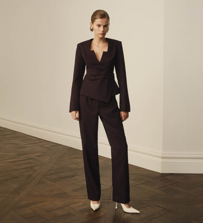 Woman wearing a dark brown suit standing in a room with wooden flooring and white walls.