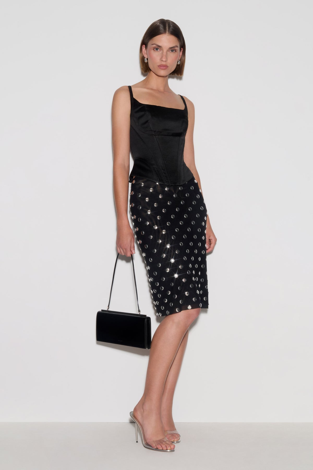 A model wearing a black satin camisole top and a black studded pencil skirt, holding a black velvet clutch bag, posed against a plain white studio background.