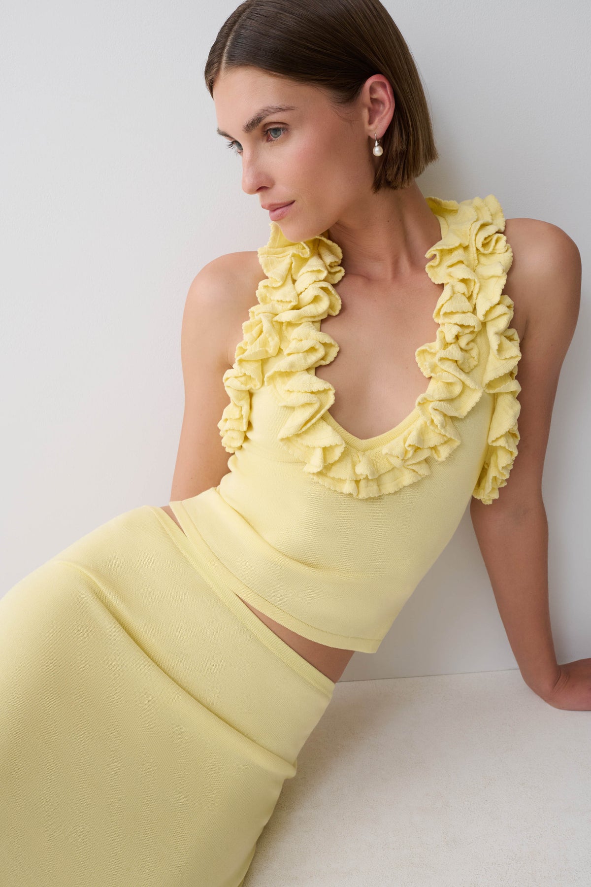A woman wearing a pale yellow ruffled halter crop top and matching high-waisted skirt, leaning against a light gray wall.