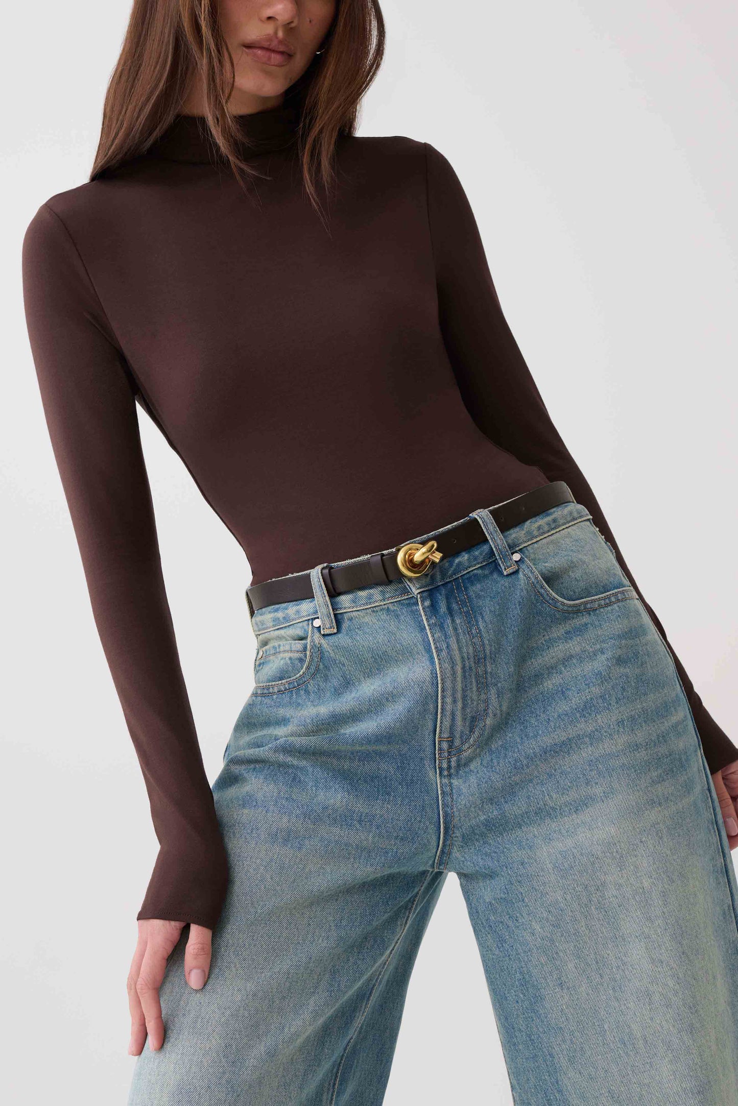 A woman wearing a fitted dark brown long-sleeve mock neck top tucked into light-wash blue jeans with a black belt featuring a gold buckle.