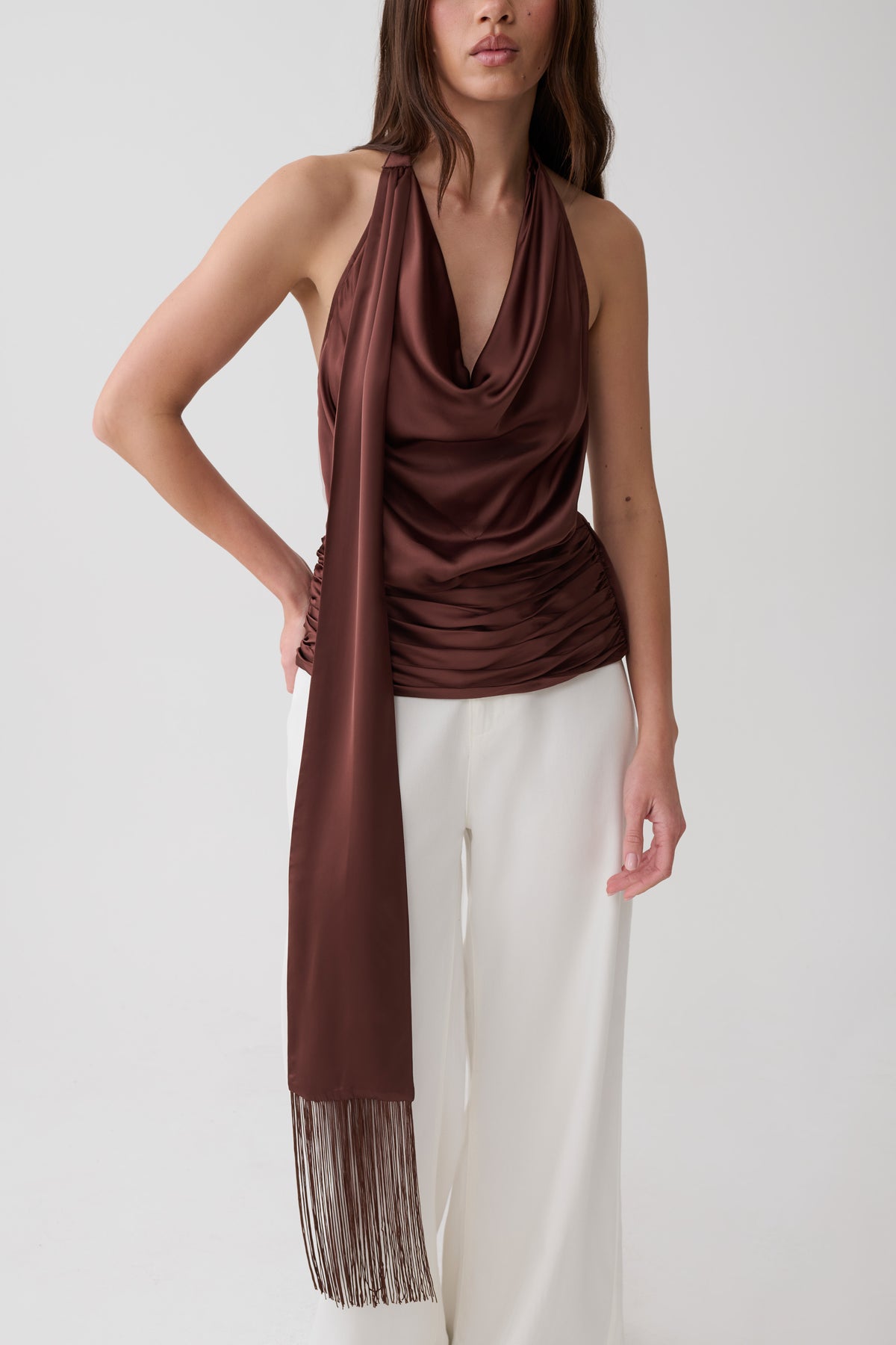A woman modeling a dark brown draped halter top with a matching long scarf draped over her shoulder, paired with white wide-leg pants, against a plain light gray studio background.