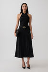 A woman modeling a sleeveless black midi dress with a halter-style neckline, a matching black belt with a gold buckle, black pointed-toe heels, and a small black clutch, photographed against a light gray studio background.