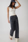 A woman modeling a pair of dark gray wide-leg jeans with contrast light stitching, styled with a cream ribbed tank top, a navy baseball cap, and white heels against a plain light gray studio background.