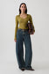 A woman stands against a light gray studio background wearing a fitted olive green long-sleeve top, wide-leg dark blue jeans with a brown belt, dark brown pointed flats, and holding a brown leather clutch.