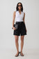 A model stands against a light gray studio background wearing a white sleeveless top, black high-waisted shorts with a black belt, black sandals, and holding a black clutch.