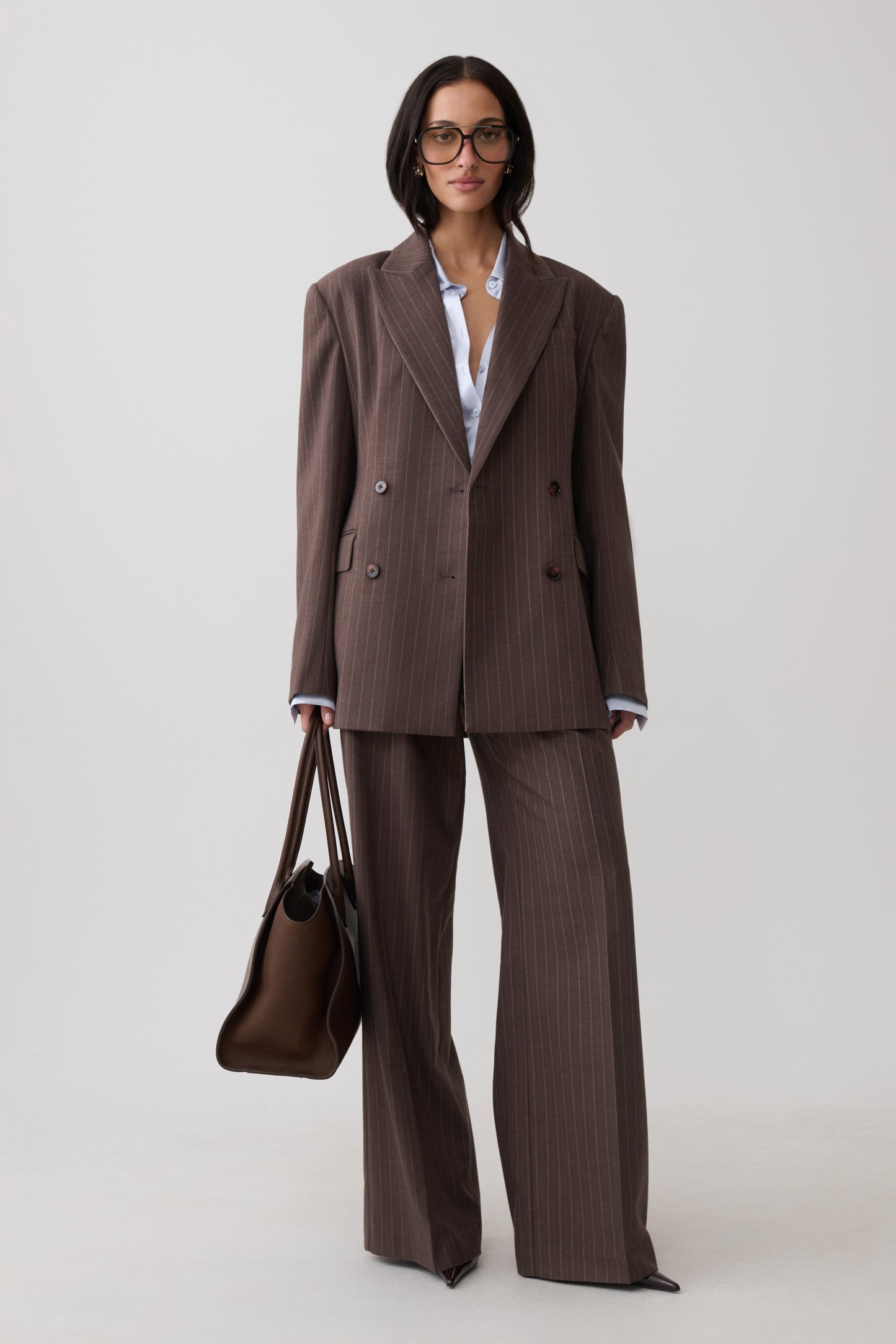 A model wearing a brown double-breasted pinstripe suit with matching wide-leg trousers, a white collared shirt, glasses, and holding a brown leather handbag against a light gray studio background.