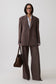A model wearing a brown double-breasted pinstripe suit with matching wide-leg trousers, a white collared shirt, glasses, and holding a brown leather handbag against a light gray studio background.