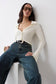 A woman wearing a fitted cream long-sleeve collared top, blue jeans, and black sunglasses leaning against a white wall on a white pedestal.