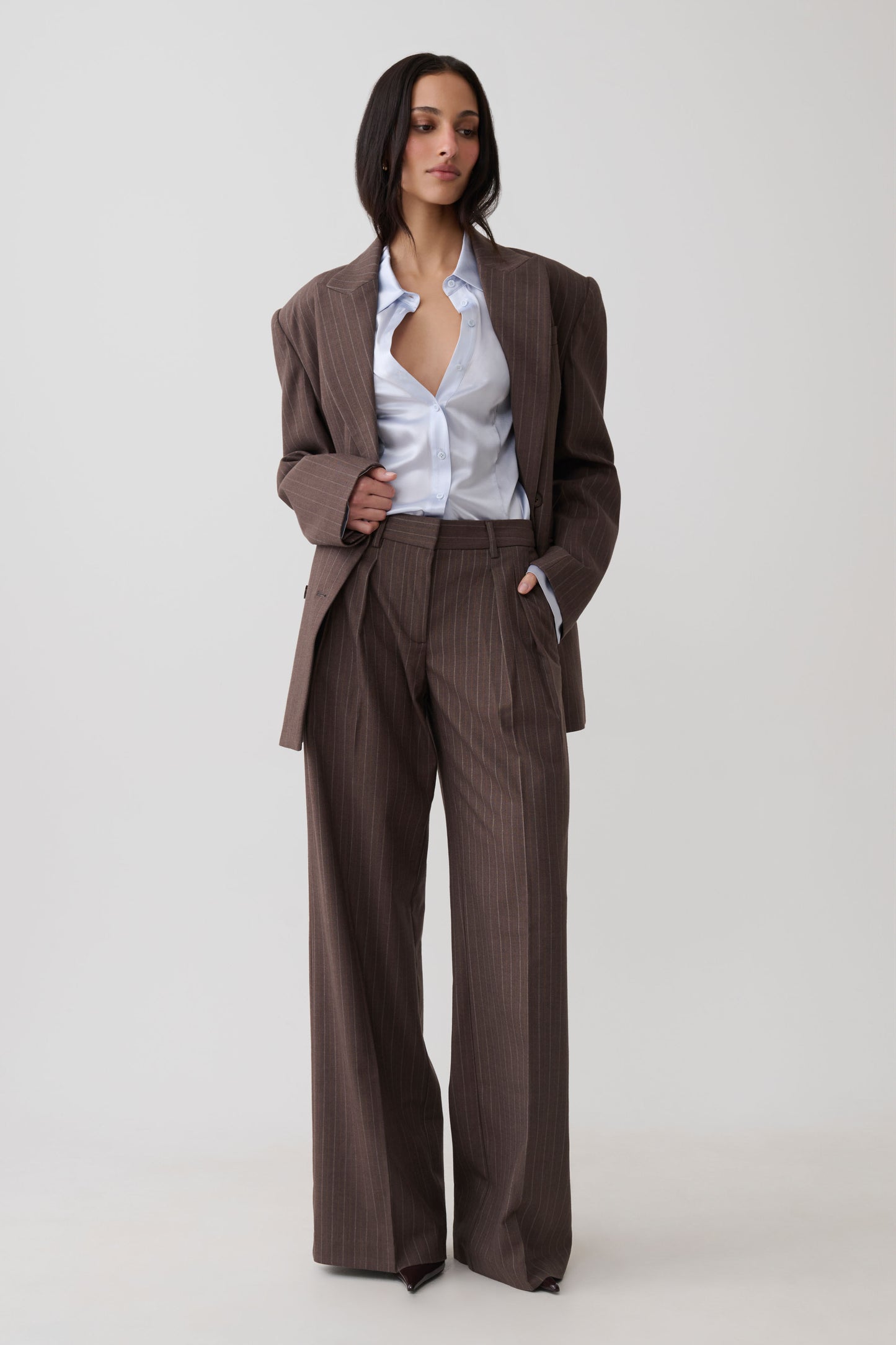 A model stands against a light gray studio background wearing a matching brown pinstripe blazer and wide-leg trousers set, styled with an open light blue button-down shirt.