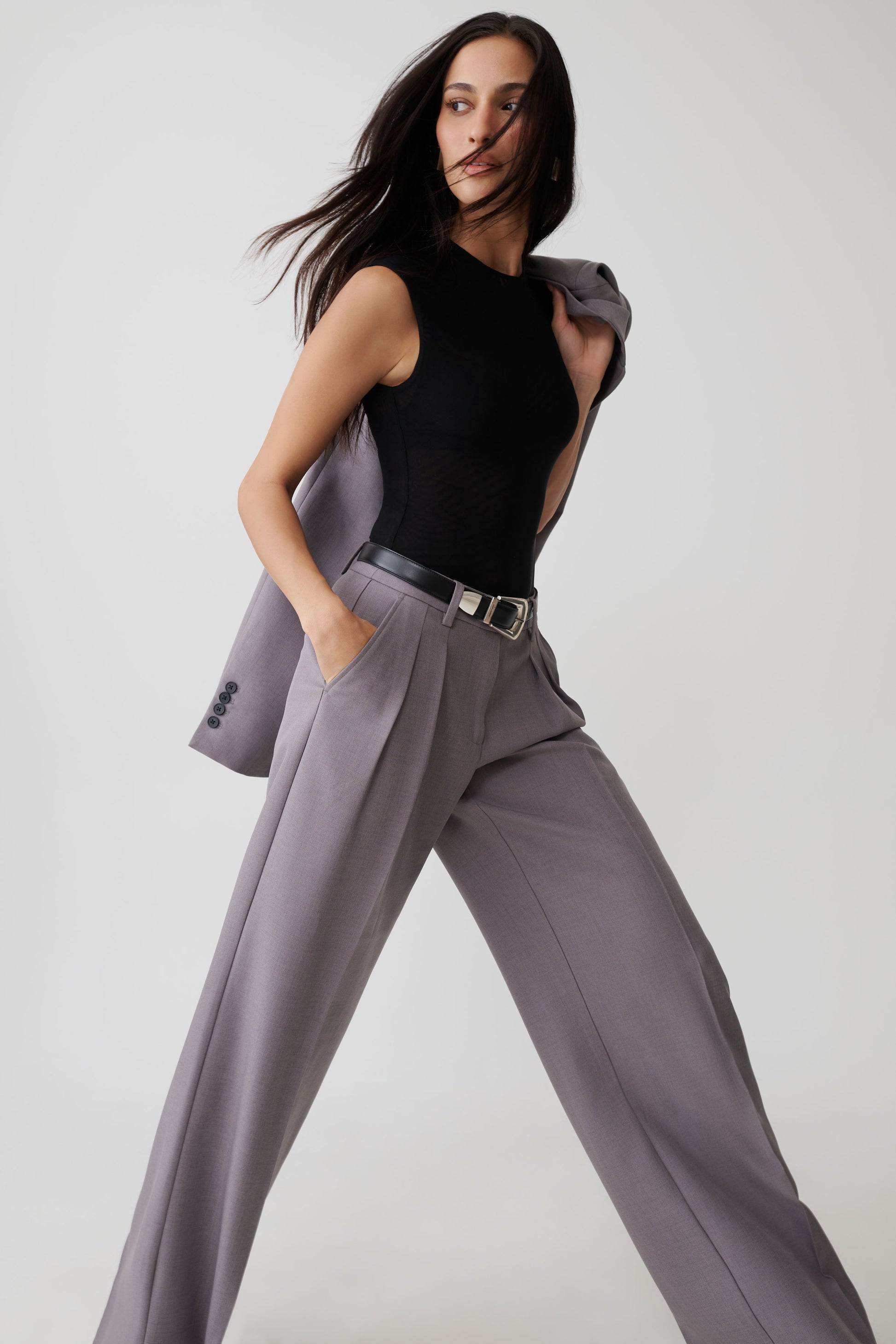 A model wearing a black sleeveless top and high-waisted, wide-leg gray trousers with a black belt, posing against a light gray studio background.