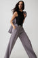 A model wearing a black sleeveless top and high-waisted, wide-leg gray trousers with a black belt, posing against a light gray studio background.