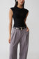 A model wearing a black sleeveless textured top and high-waisted gray pleated trousers with a black belt featuring a silver buckle, shot against a light gray studio background.