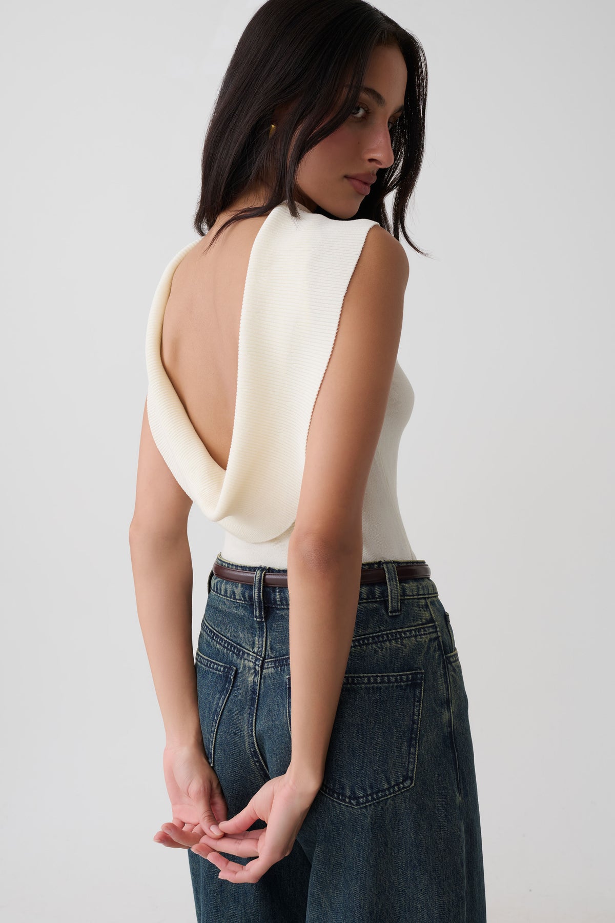A woman wearing a cream-colored sleeveless top with a dramatic draped open back, paired with high-waisted dark blue jeans, photographed against a plain light gray studio background.