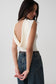 A woman wearing a cream-colored sleeveless top with a dramatic draped open back, paired with high-waisted dark blue jeans, photographed against a plain light gray studio background.