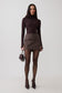 A model stands against a light gray studio background wearing a dark brown mock-neck long-sleeve top, a matching brown pleated wrap mini skirt, sheer black tights, and dark pointed-toe heels, while holding a small dark brown handbag.
