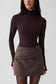 A model wearing a dark brown mock neck long-sleeve top and a brown pinstriped wrap mini skirt, photographed against a light gray studio background.