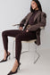 A woman models a dark brown pinstripe blazer, matching dark brown mock-neck top, and dark brown leggings while seated on a modern chair with a silver metal frame.