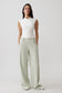 A woman modeling a white sleeveless wrap-style top and light sage green wide-leg trousers against a plain light gray studio background.