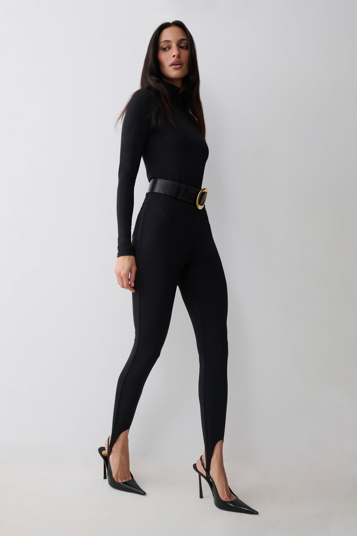 A woman modeling a black long-sleeve bodysuit with black stirrup leggings  with a wide black belt featuring a gold oval buckle, paired with black pointed-toe stiletto heels, against a light gray studio background.