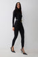 A woman modeling a black long-sleeve bodysuit with black stirrup leggings  with a wide black belt featuring a gold oval buckle, paired with black pointed-toe stiletto heels, against a light gray studio background.