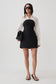 A model stands against a light gray studio background wearing a black strapless mini dress with a white long-sleeve collared shirt layered over it, black sunglasses, black sandals, and holding a black handbag.