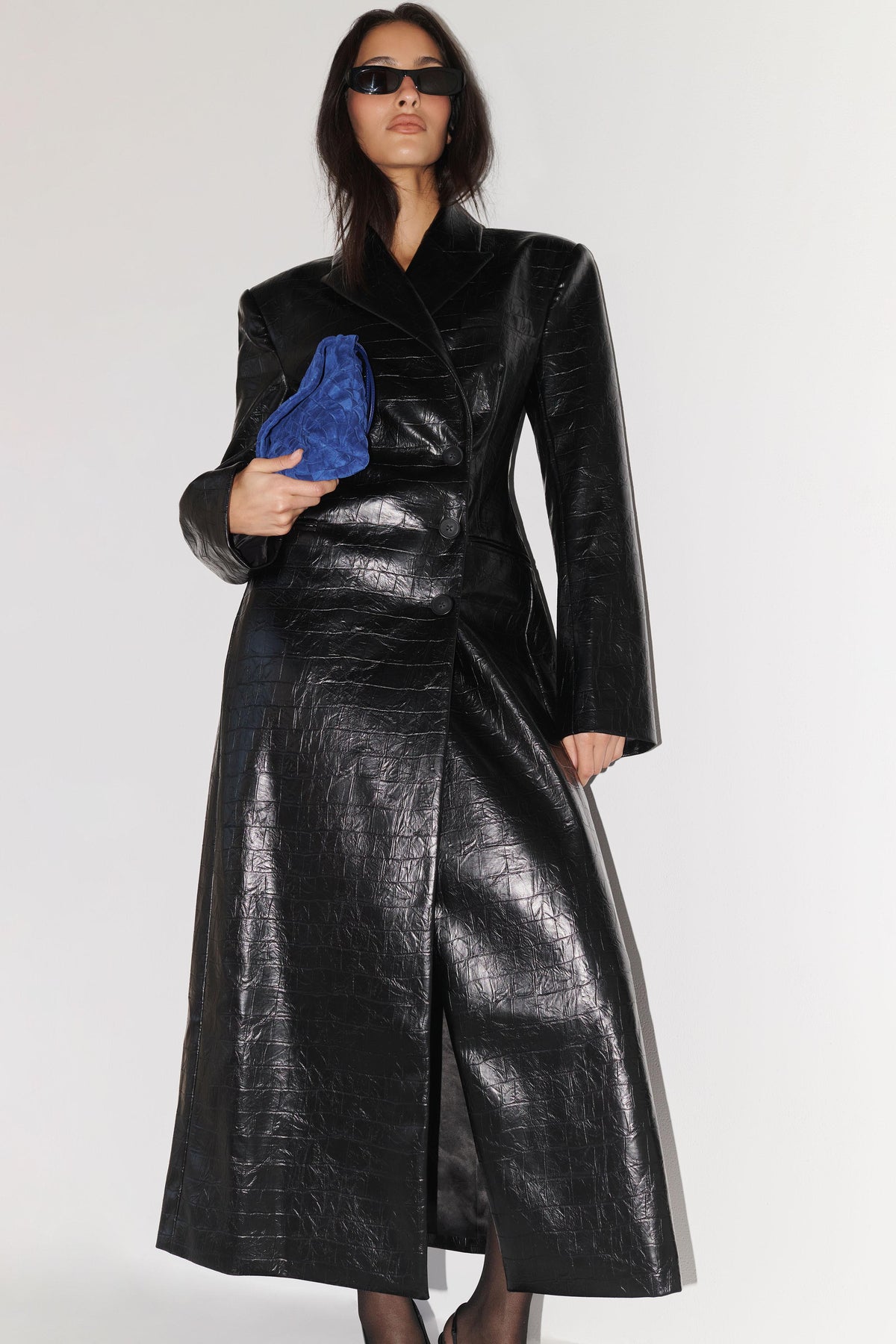 A model wearing a long, structured black faux-leather coat with a glossy, embossed texture, holding a blue velvet clutch against a plain light gray studio background.