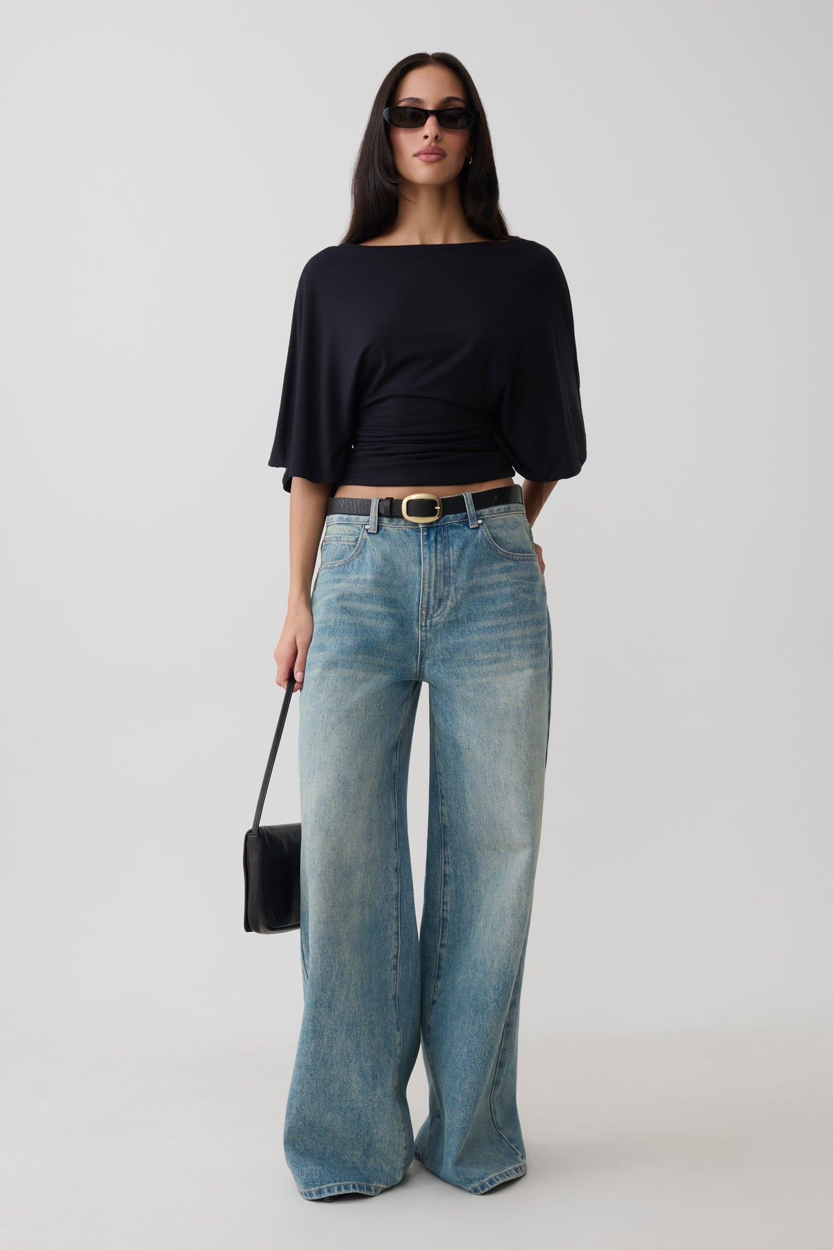 A woman modeling a black boat-neck top with elbow-length sleeves and light-wash wide-leg jeans, styled with a black belt, sunglasses, and a small black handbag against a light gray studio background.