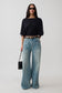 A woman modeling a black boat-neck top with elbow-length sleeves and light-wash wide-leg jeans, styled with a black belt, sunglasses, and a small black handbag against a light gray studio background.