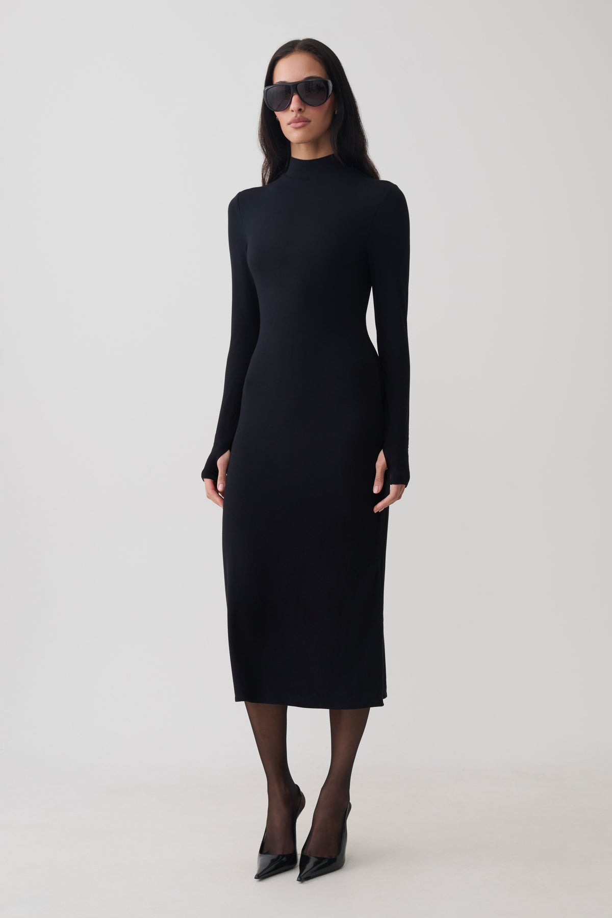 A woman modeling a fitted black long-sleeve mock-neck midi dress, styled with black sunglasses, sheer black tights, and black pointed-toe heels against a plain light gray studio background.