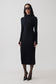 A woman modeling a fitted black long-sleeve mock-neck midi dress, styled with black sunglasses, sheer black tights, and black pointed-toe heels against a plain light gray studio background.