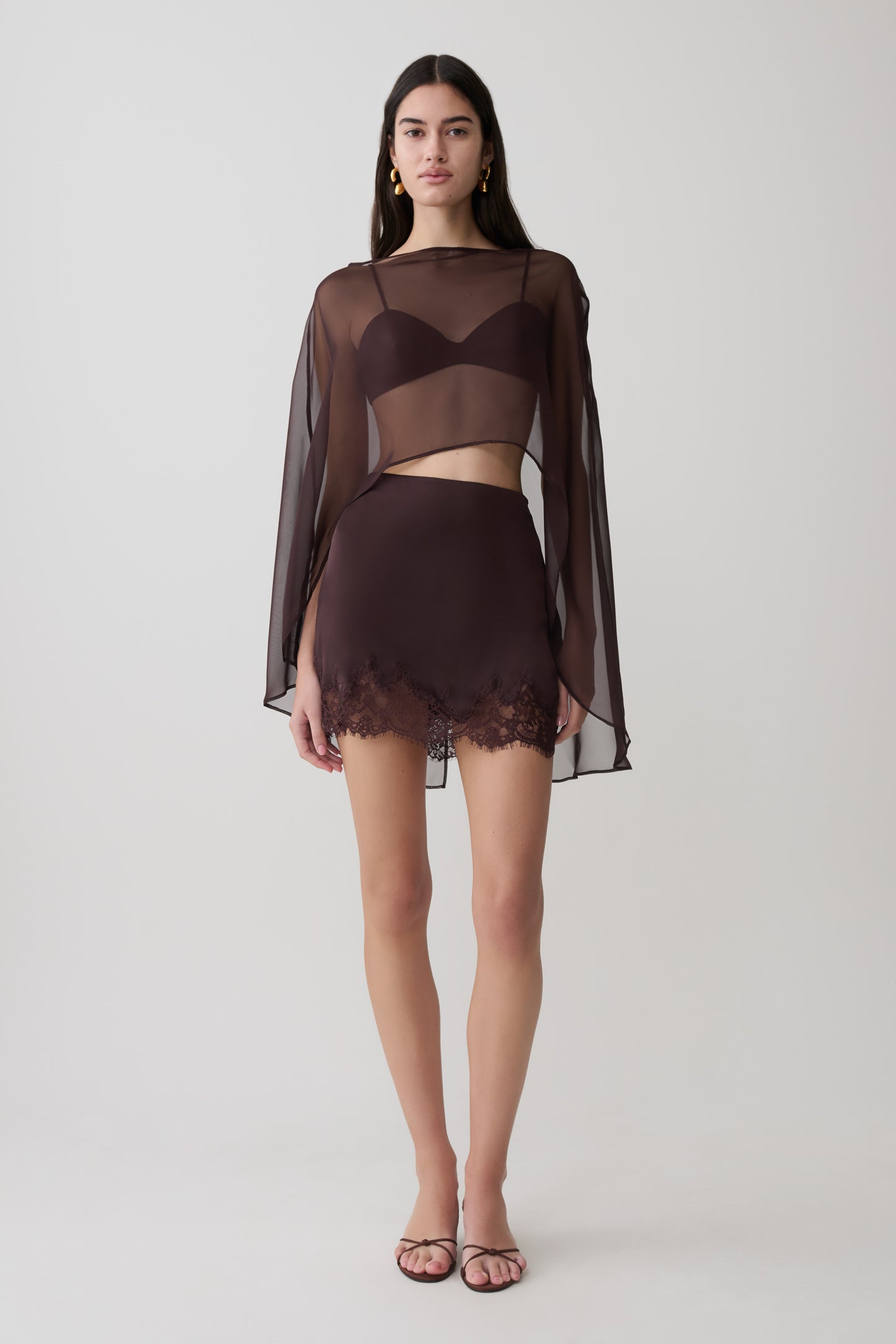 Woman wearing brown bralette with chiffon cape with matching lace and satin mini skirt.