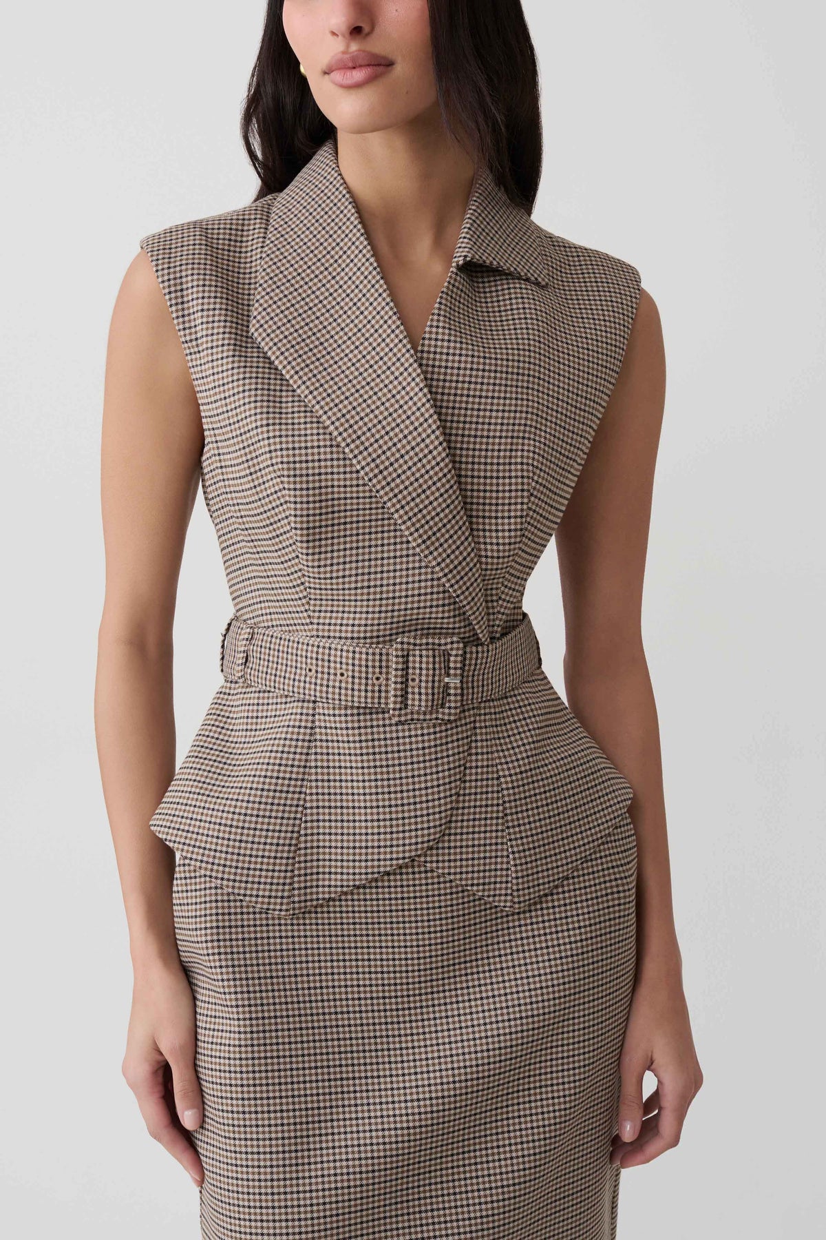 A woman wearing a sleeveless belted wrap dress with a fine brown and beige houndstooth pattern, shown from the neck down against a light gray studio background.