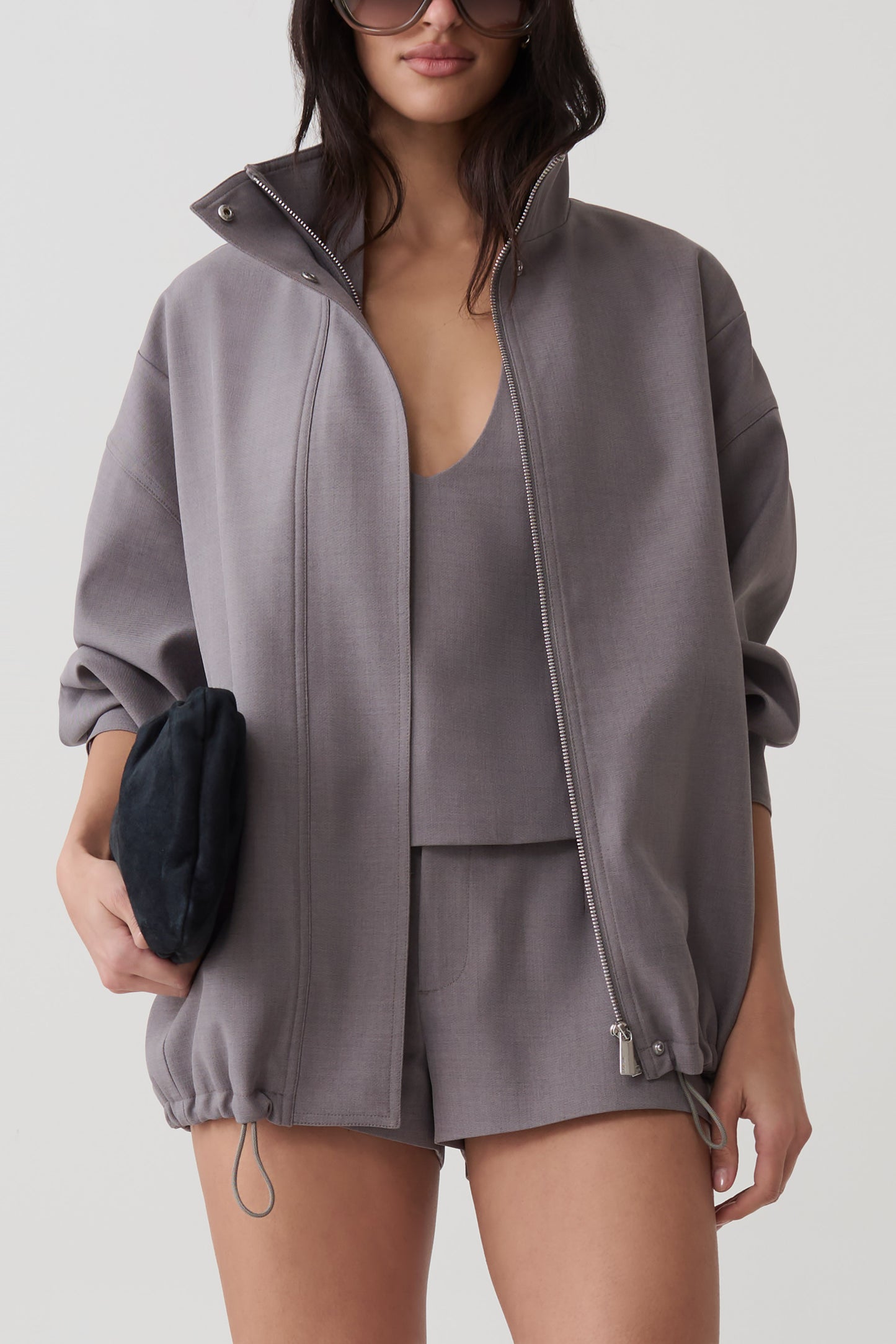 A woman models an oversized gray zip-up jacket with a stand collar, paired with matching shorts and a matching camisole. She holds a small black clutch against a clean, light gray studio background.