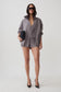 A model stands against a light gray studio background wearing a coordinated gray outfit consisting of an oversized zip-front jacket, matching top, and shorts. She is also wearing black pointed heels and holding a small dark clutch.