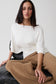 A woman wearing a white draped three-quarter sleeve top and tan wide-leg trousers, seated against a light gray background.