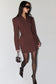 A woman modeling a dark brown blazer dress with structured shoulders, paired with black tights, pointed-toe heels, and a small matching handbag against a plain light gray background.