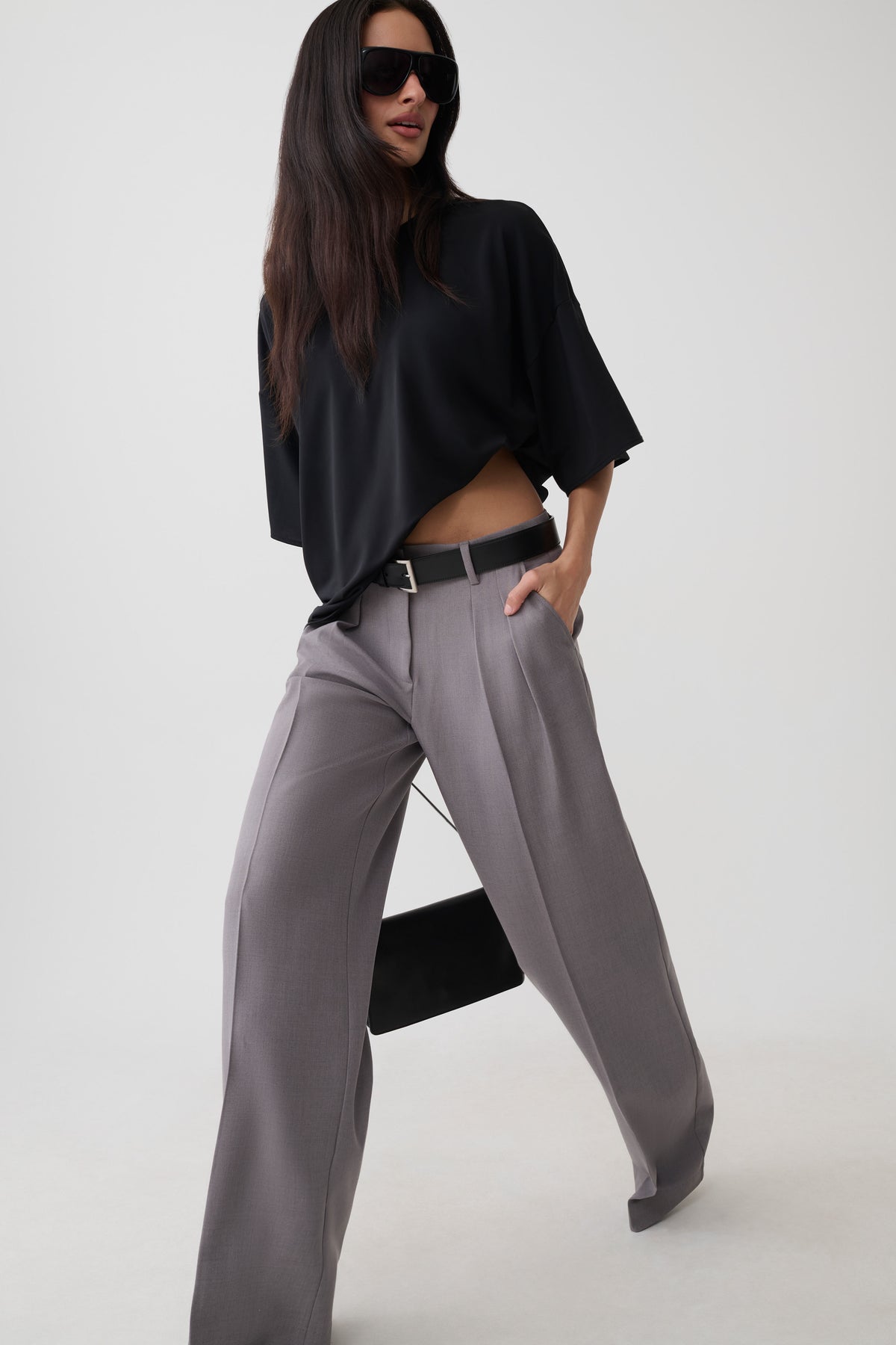 A model wearing a black short-sleeve crop top and high-waisted, wide-leg gray trousers with a black belt, posing against a light gray studio background.