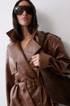 A woman wearing a brown leather trench coat with a matching belt and large dark sunglasses, holding a dark brown shoulder bag against a plain light gray background.